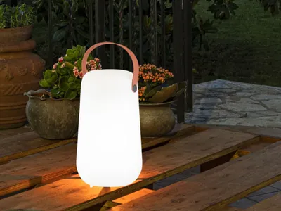 A white lantern with a brown handle sits on a wooden table outdoors at dusk, with plants in pots in the background.