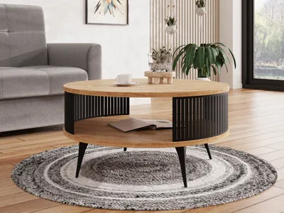 A round coffee table with a wooden top and black metal slatted sides is placed on a circular rug in a living room setting. A grey sofa is visible to the left, and a large window with greenery outside is to the right.