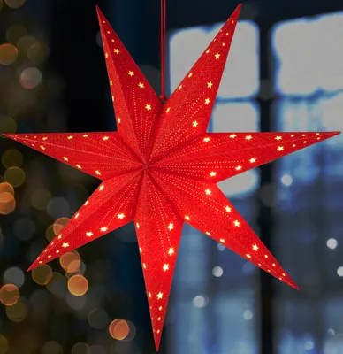 A red star-shaped lantern with small yellow stars is hanging in front of a blurred background of lights and a window.