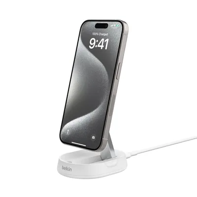 A white wireless charging stand with a silver iPhone on it, plugged in with a white USB cable.