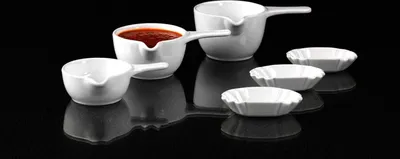 A set of white ceramic dipping bowls and sauce boats are arranged on a reflective black surface.