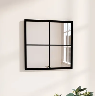 A square mirror with a black frame, divided into four panes by thin black lines, is mounted on a light-colored wall. The mirror reflects a window with natural light coming through.