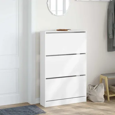 A white shoe cabinet with three flip-down drawers is shown in a hallway next to a white door. A grey rug is on the floor in front of the cabinet, and a grey tote bag and a wooden bench are to the right.