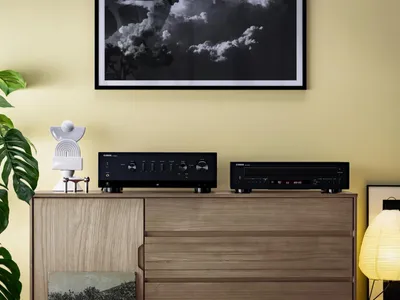 Two black audio components are placed on a wooden dresser, with a large framed black and white photograph hanging on the yellow wall above.