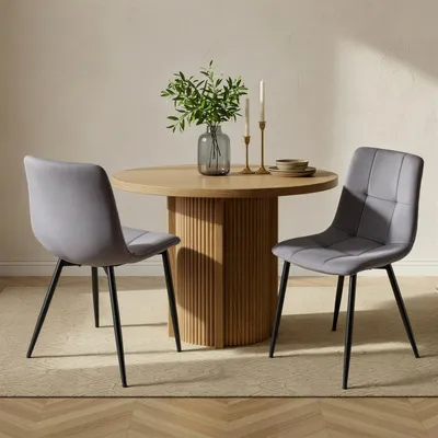 Two gray upholstered dining chairs are placed around a round wooden dining table with a fluted pedestal base. A vase with greenery and two candlesticks are on the table.