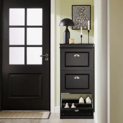 A black shoe cabinet with three flip-down drawers and open shelves is placed next to a black door in a hallway.