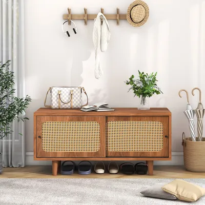 An entryway scene featuring a wooden shoe bench with rattan doors, decorated with a handbag, books, a plant, and umbrellas in a basket.