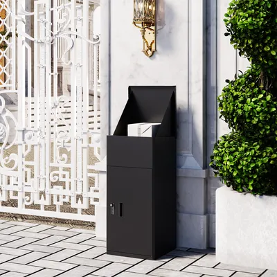 A black parcel mailbox is placed on a paved walkway next to a white ornate gate and a green bush.