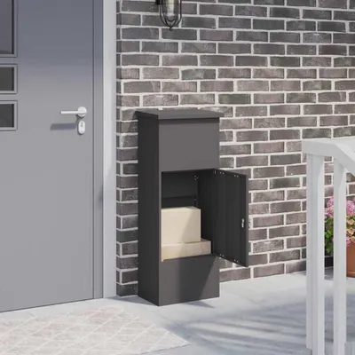A dark gray parcel drop box with its lower door open, revealing two cardboard boxes inside, is placed on a concrete patio next to a gray door and a brick wall.