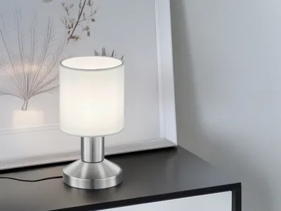 A modern table lamp with a white fabric shade and a brushed silver base is placed on a dark surface next to a framed picture.