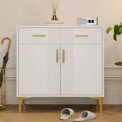A white sideboard with gold legs and handles, featuring two drawers and two doors, is shown in a room setting. An umbrella stand with an umbrella is to the left, and slippers are on the floor in front. A diffuser and a small radio are on top.