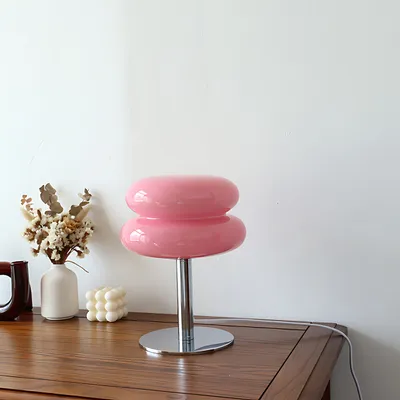 A pink glass table lamp with a chrome base sits on a wooden dresser next to a vase of dried flowers and a white cube-shaped candle.