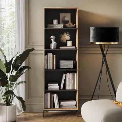 A tall, narrow bookshelf filled with books and decorative items, placed in a well-lit room next to a large plant and a floor lamp.