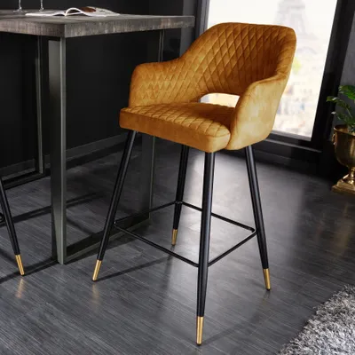 A mustard yellow velvet bar stool with diamond quilting and gold foot caps is shown next to a dark wood and metal bar table.