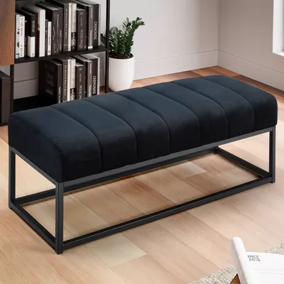 A black velvet bench with a metal frame is placed in a room with a bookshelf and a plant.