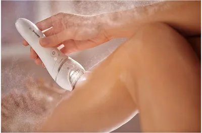 A person is using a white epilator on their leg, with a fine mist of water in the air.