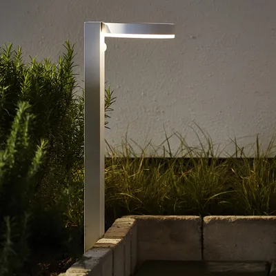 A modern outdoor lamp is shown in a garden setting at dusk.