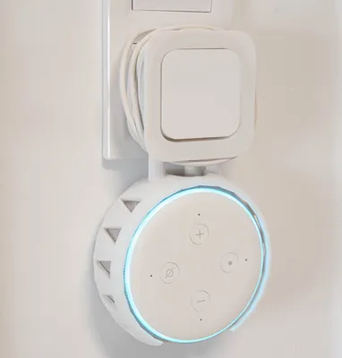 A white wall mount for an Amazon Alexa Echo Dot 3rd generation is plugged into a wall socket.