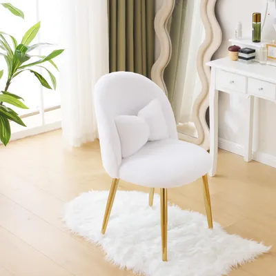 A white plush chair with gold legs and a heart-shaped pillow is placed on a white rug in a room with a large window and a vanity table in the background.