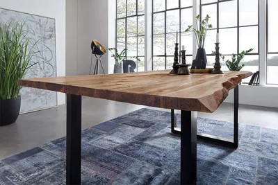 A dining table with a live edge top and black U-shaped legs, set in a modern dining room with large windows overlooking a body of water.