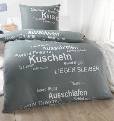 A grey duvet cover and pillowcase with white text printed on them, laid out on a wooden floor with a white blanket in the background.