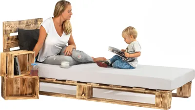 A woman and a child are sitting on a bed made of wooden pallets. The woman is reading a book, and the child is looking at a tablet.