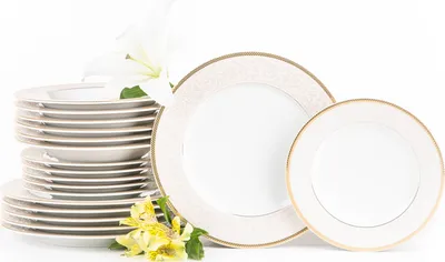 A stack of white dinner plates with gold trim and two decorative plates, accompanied by white and yellow flowers, on a white background.