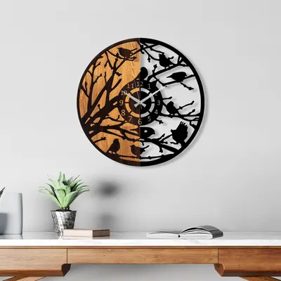A wall clock with a bird and tree design is displayed on a white wall above a wooden desk.