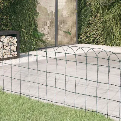 A garden scene with a green wire fence in the foreground, a paved area with white gravel, and lush green plants in the background.