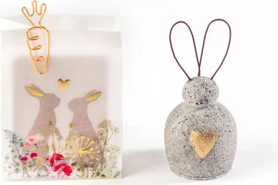 Two Easter-themed decorative items: a white paper bag with a carrot clip and bunny graphics, and a gray ceramic bunny bell with a gold heart.