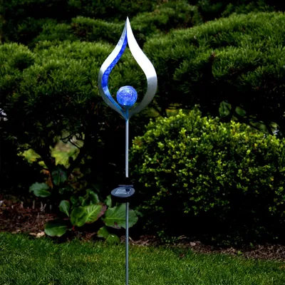 A solar-powered garden stake with a flame-shaped metal design and a blue glass ball, set in a garden with lush green bushes and grass.
