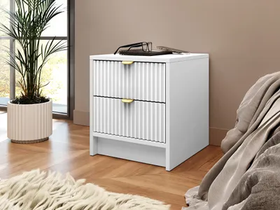 A white nightstand with two drawers and gold handles is placed next to a bed. A plant in a ribbed pot is on the left, and a fluffy rug is in the foreground.