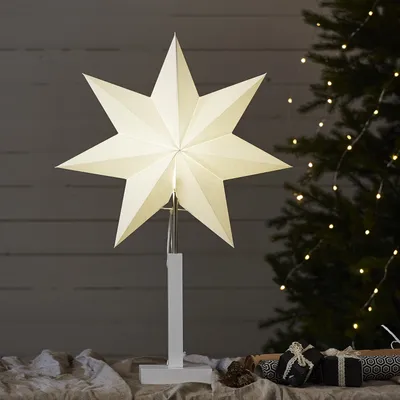 A white star-shaped lamp on a stand, placed on a table with gifts and a Christmas tree in the background.