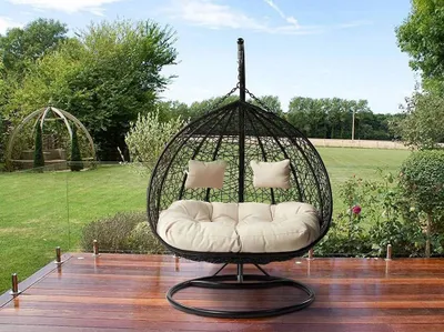 A hanging egg chair with cushions is placed on a wooden deck in a garden setting.