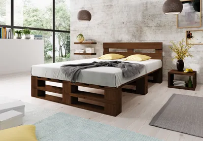 A bedroom scene featuring a wooden pallet bed with a headboard, pillows, and a blanket. The room has a textured wall, shelves with decor, a bedside table, and a large window with a view of greenery.
