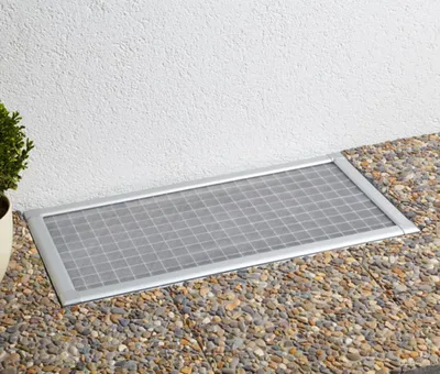 A rectangular metal grate cover with a grid pattern is placed on a gravel path next to a white textured wall.