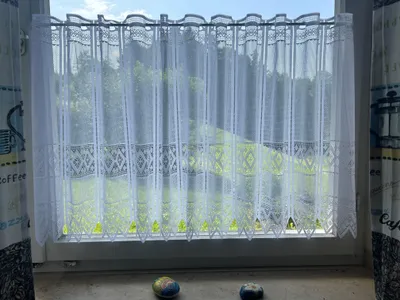 A white lace curtain is hung on a window, with a view of greenery and sky behind it. Two decorative stones are on the windowsill.
