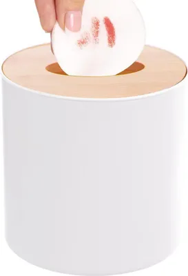 A hand is shown dropping a used cotton pad into a white cosmetic bin with a wooden lid.