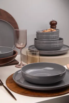 A collection of grey ceramic tableware, including bowls and plates, is arranged on a white surface. A pink wine glass and a wooden pepper mill are also visible.