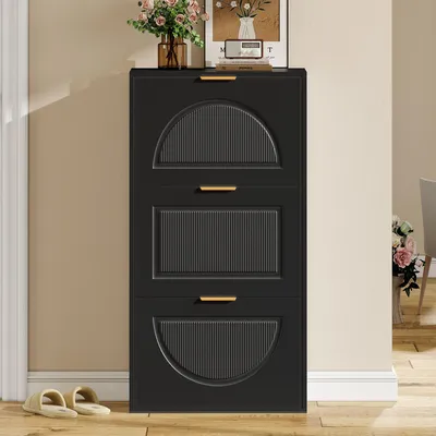 A black shoe cabinet with three flip-down doors, decorated with fluted panels and gold handles, stands against a light beige wall. A vase of flowers and a framed picture are on top of the cabinet. A pair of slippers rests on the wooden floor in front of th