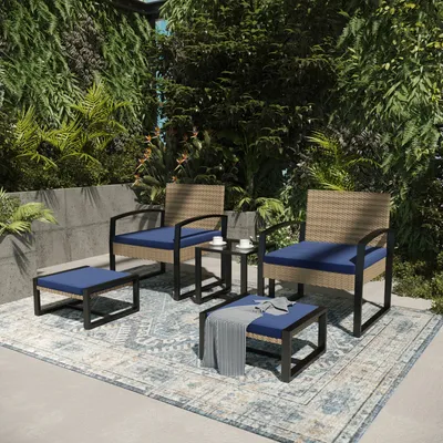 An outdoor patio set with chairs, ottomans, and a table, surrounded by lush greenery and a patterned rug.