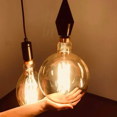 A hand is holding a large, clear glass light bulb with an amber tint, showcasing its size. Another similar light bulb hangs in the background.