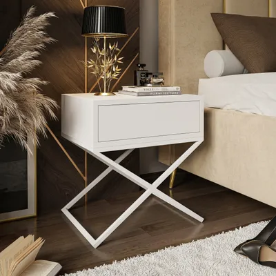 A white nightstand with a drawer is placed next to a bed in a bedroom. A lamp, books, and decorative items are on top of the nightstand. Dried plants are on the left, and a rug is on the floor.