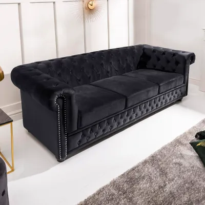 A black velvet Chesterfield sofa is shown in a well-lit room with a white paneled wall and a grey rug.