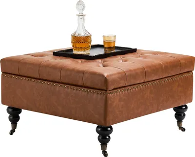 A square leather ottoman with tufted top and decorative nailhead trim sits on black legs with casters. A decanter and glass of amber liquid are on a black tray on top of the ottoman.