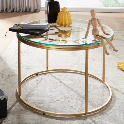 A round glass coffee table with a gold metal frame, decorated with a wooden mannequin, a yellow vase, and a stack of books.