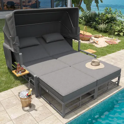 A double lounger with a canopy and side tables is set up on a patio next to a swimming pool, with a picnic setup in the background.