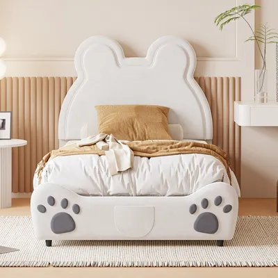 A white children's bed with a bear-ear shaped headboard and paw print details on the footboard, set in a bedroom with a beige fluted wall panel and a small white side table.
