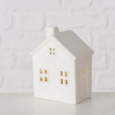 A white ceramic house-shaped decoration with cut-out windows and a chimney, placed on a white surface against a white brick wall.