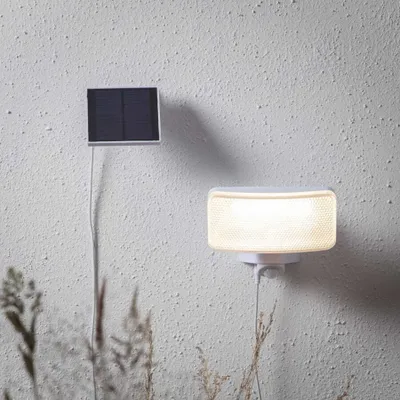 A solar-powered LED light and its solar panel are mounted on a textured wall, with dry grass in the foreground.